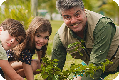Homem com colete de biólogo e duas crianças plantando uma muda de árvore em um gramado.