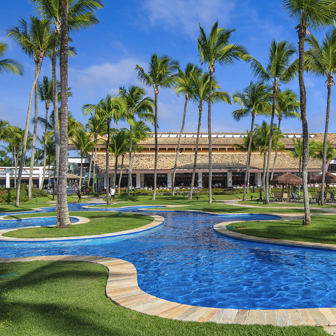 Piscina sinuosa rodeada por coqueiros e jardins tropicais em resort à beira-mar sob céu azul ensolarado.