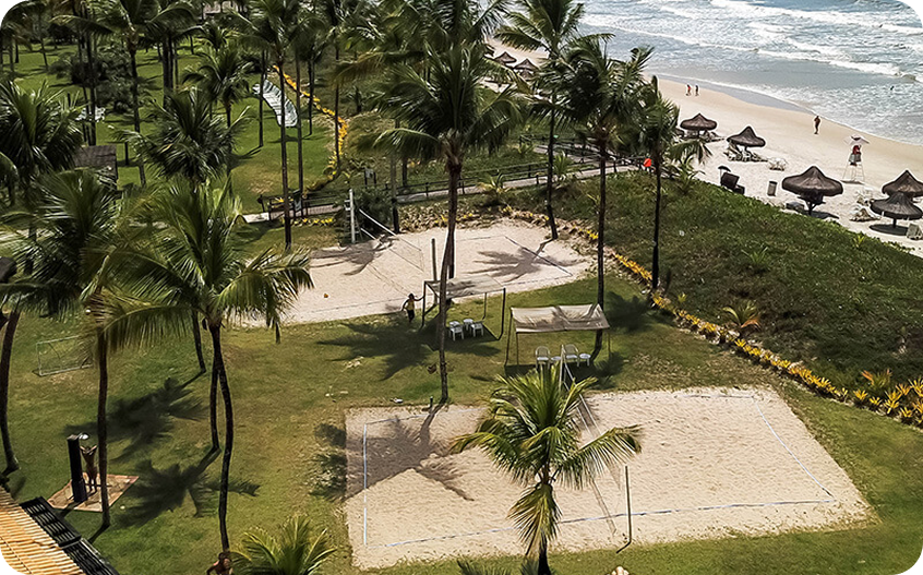 Quadras de vôlei de praia cercadas por coqueiros em resort à beira-mar, com vista para o oceano e estrutura de lazer tropical