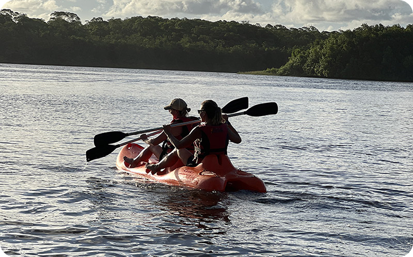 Duas pessoas remando em caiaque vermelho sobre lago tranquilo cercado por vegetação tropical ao entardecer